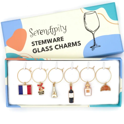 Six small wine charms attached to key rings, displayed in a box. The designs are of the French flag, the Moulin Rouge, the Eiffel tower, a wine bottle, a perfume bottle, and a croissant. Above the charms, text says, "Serendipity Stemware Glass Charms."