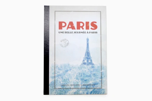 A notebook with a blue retro Eiffel Tower illustration on its cover. The cover bottom reads "Une belle journée à Paris" (a beautiful day in Paris).
