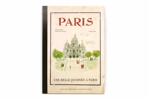 A notebook with a retro Montmartre illustration on its cover. The cover bottom reads "Une belle journée à Paris" (a beautiful day in Paris).