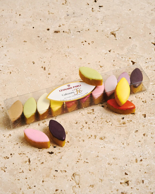 A clear 65 gram box of Léonard Parli mini calissons on a stone countertop. The ten calissons are each a different pastel color.