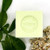 A 100 gram light yellow colored cube of Senteurs de France's verbena-scented Marseille cube. The cube sits atop leaves and flowers.