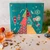 A square teal "duo" advent calendar with organic tea from Provence d'Antan. The calendar is in two clear segments and the front features an abstract Christmas tree with segments of various patterns. The calendar sits on a table.