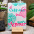 A card from L'Atelier Letterpress that reads, "Cultivons notre amour" in pink and cyan lettering in front of an illustration of a pink watering can and cyan bushes. The card sits on a display stand.