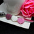 Three Ercus mini heart sugar pieces in pink, white, and purple, in a line on a plate with a tea cup, spoon and pink rose.