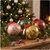 Four Christmas balls with strings in different colors (red, green, golden, and pink) with a Christmas tree in the background.