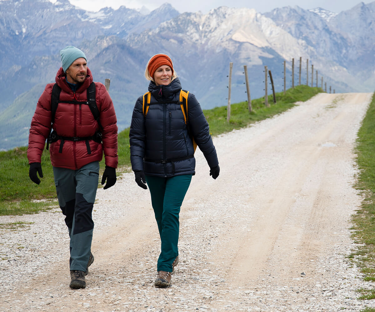 Man and woman walking on mountain path, with snowy mountains behind, wearing Insulated Eos Jackets.