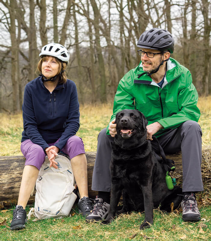 Two people sat on a fallen tree, taking a break from cycling with their dog