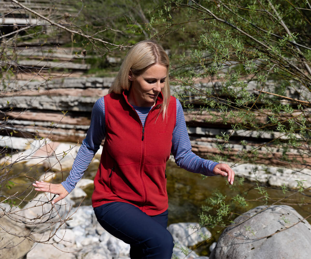 Woman walking over rocks, beside stream, wearing New Season Microgrid Fleece Gilet.