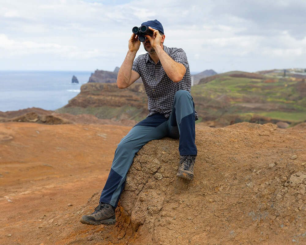 Man sitting on sandy rocks looking through binoculars, wear Walking Trousers, Walking Boots, a Shirt and a Cap.