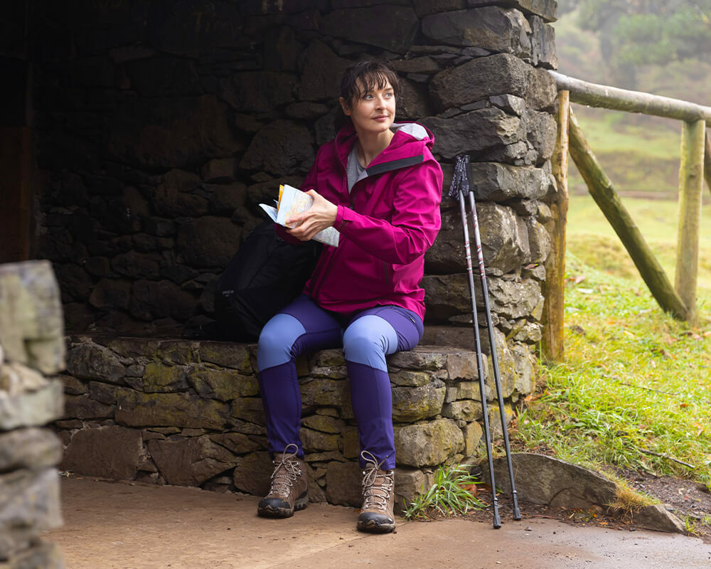Woman sat in a brick shelter reading a map, wear Waterproof Jacket, Walking trousers and Walking Boots, with Walking Poles propped up.