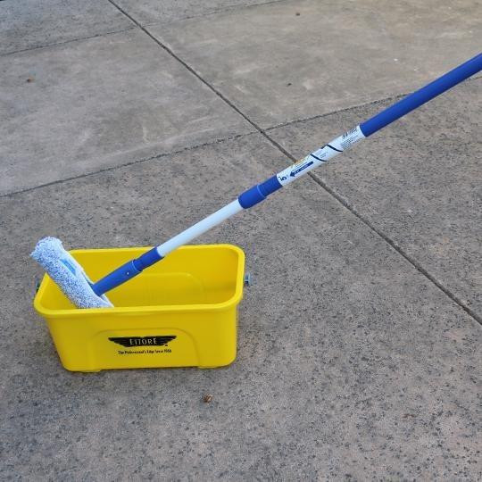 Ettore Mighty Window Washers, window cleaning kit with a blue and white extendable pole and a microfiber cleaning head, placed on a yellow cleaning bucket on a concrete surface.