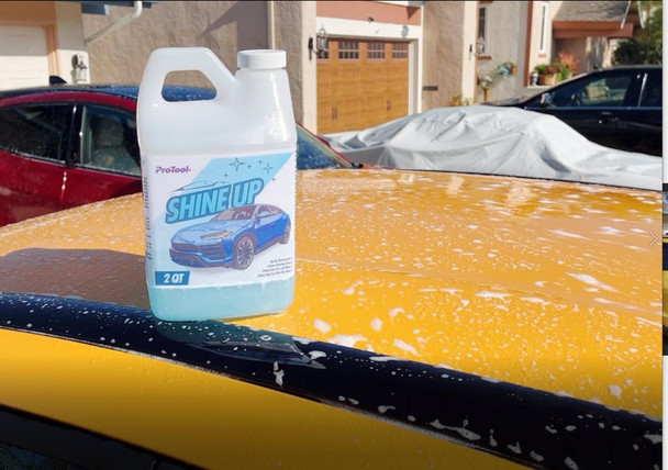 ProTool Foaming Hose End Sprayer, a 2-quart white plastic bottle with a label featuring a blue car and the text 'Shine Up' on it, placed on a yellow car hood covered with foam.