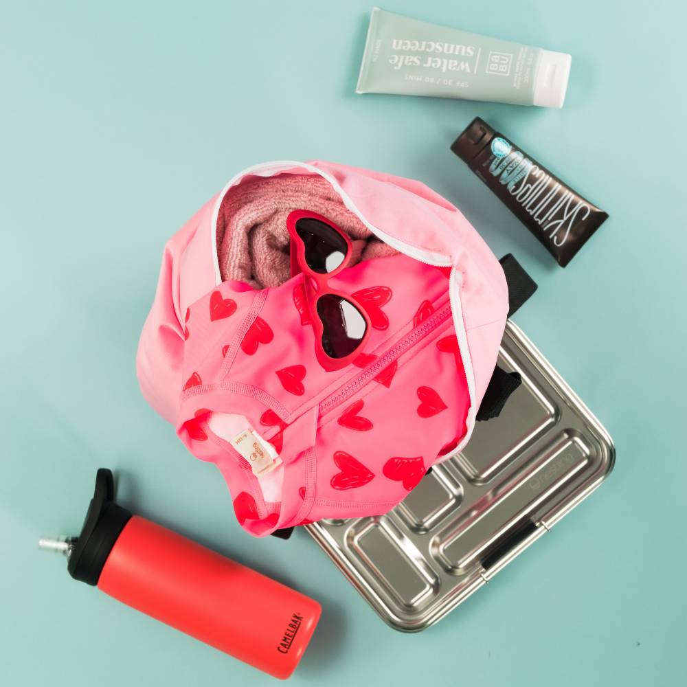 Flat lay - sunscreen, double pocket wetbag, drink bottle, lunchbox, sunglasses, swimwear