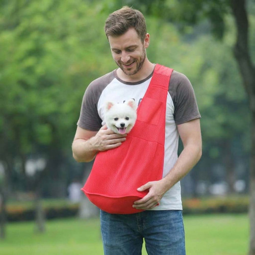 Man wearing a red mesh dog sling, carrying a smiling white Pomeranian dog for a hands-free walk in the park.