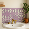 Lavender daisy tiles behind a white sink with gold faucet and a potted plant.