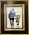 Hopes For The Cup is an original oil painting with a football theme by Alexander Millar. This image show the artwork framed in a steely blue/grey frame with burnished gold mouldings. The adult is in a shabby, blue work suit with a flat cap, typical of the attire of working men from the mid 20th century. The adult is carrying an old, leather football under his left arm and has his right arm on the shoulders of the child. The child is wearing a dark green balaclava and  a brown wool coat or a duffle coat. The child is pointing up to the sky, obviously in excited conversation with the adult about his football team's performance.