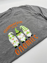 Gray shirt with a graphic of three gnomes with long white beards, each dressed in a different Pattern with a matching hat, green and white buffalo plaid, green and white polka dot, and green and white houndstooth, surrounded by a logo that reads "Shamrockin with my Gnomies" in Orange Soda EasyWeed Heat Transfer Material shadowed with Gold EasyWeed Heat Transfer Material.