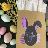 A Brown paper bag with handles with a bunny image created with Blackboard Heat Transfer Vinyl with the name "Taylor" written on it with chalk. The inside of the bunny's ears are colored with neon purple Glitter HTV and it is framed with a stylized egg on either side in Strip Flock Pro.