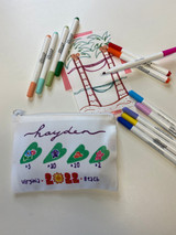 Image shows a small pouch bag in the foreground with four green hearts across the middle with shells and flowers inside of them. Above the hearts reads the name "Hayden" and across the bottom reads "Virginia 2022 Beach" - slightly overlapping a pastel colored tic tac toe grid depicted by 2 palm trees connected by a hammock and waves. Primary and pastel markers surround both.