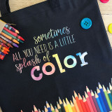 Image shows a black tote bag with a logo that reads "Sometimes all you need is a little splash of color" with a rainbow watercolor appearance. There is a blue button the top right of the bag, a box of crayons sitting on the top left and the bottom of the bag is lined with a color pencil graphic that are in order based on the color wheel.