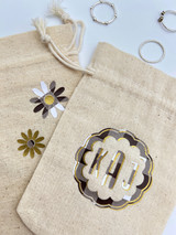 Image of two beige cinch bags with the logo of a “scalloped circle” and the initials “KAJ” using brown Buffalo Plaid and logo of “daisy-like flowers” using the brown Buffalo Plaid. Both logos have specs of EasyWeed Gold incorporated.