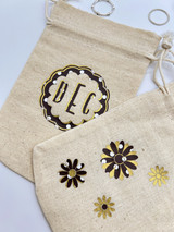 Image of two beige cinch bags with the logo of a “scalloped circle” and the initials “DEC” using brown Polka Dot and logo of “daisy-like flowers” using the brown Polka Dot Pattern. Both logos have specs of EasyWeed Gold incorporated.