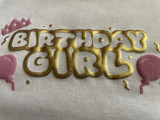 White shirt with a logo that reads "Birthday Girl" in hollowed Gold Puff Metallic Heat Transfer Vinyl. Surrounding the logo, there are confetti and streamer pieces in Gold Puff Metallic, balloons on either side in Pink Puff Heat Transfer Vinyl and a crown in Pink Puff Heat Transfer Vinyl atop the B in Birthday.