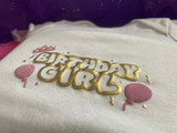 White shirt with a logo that reads "Birthday Girl" in hollowed Gold Puff Metallic Heat Transfer Vinyl. Surrounding the logo, there are confetti and streamer pieces in Gold Puff Metallic, balloons on either side in Pink Puff Heat Transfer Vinyl and a crown in Pink Puff Heat Transfer Vinyl atop the B in Birthday.
