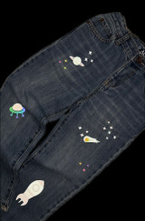 Image of jeans with space related graphics including stars, a planet, comet, rocket, and spaceship made with Dark Gray EasyReflective Heat Transfer Vinyl, Silver EasyReflective Heat Transfer Vinyl, Aqua EasyReflective Heat Transfer Vinyl, and Rainbow Black EasyReflective Heat Transfer Vinyl with light shining on them, appearing illuminated.