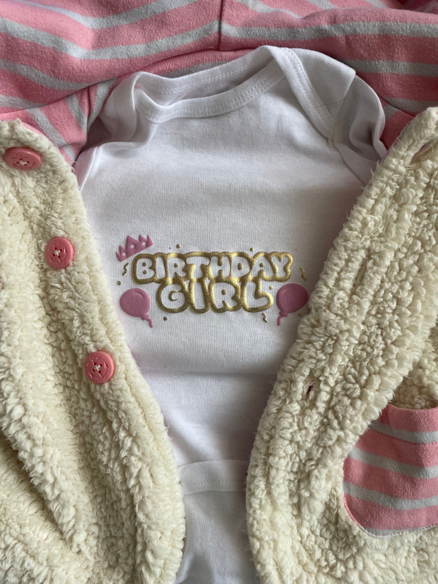 Image of a white onesie with a logo that reads "Birthday Girl" in Gold Puff Metallic Heat Transfer Vinyl. Surrounding the logo, there are confetti and streamer pieces in Gold Puff Metallic, balloons on either side in Pink Puff Heat Transfer Vinyl and a crown in Pink Puff Heat Transfer Vinyl atop the B in Birthday.