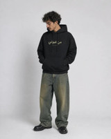Model wearing a black pullover hoodie with small olive-green Arabic and English text on the chest, front view