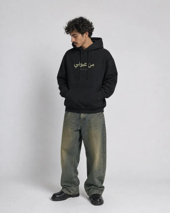 Model wearing a black pullover hoodie with small olive-green Arabic and English text on the chest, front view