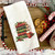 Close-up of All Booked for Christmas dish towel — rich jewel-toned stacked books with holly berries, candy cane and steaming cocoa sublimated on white waffle weave