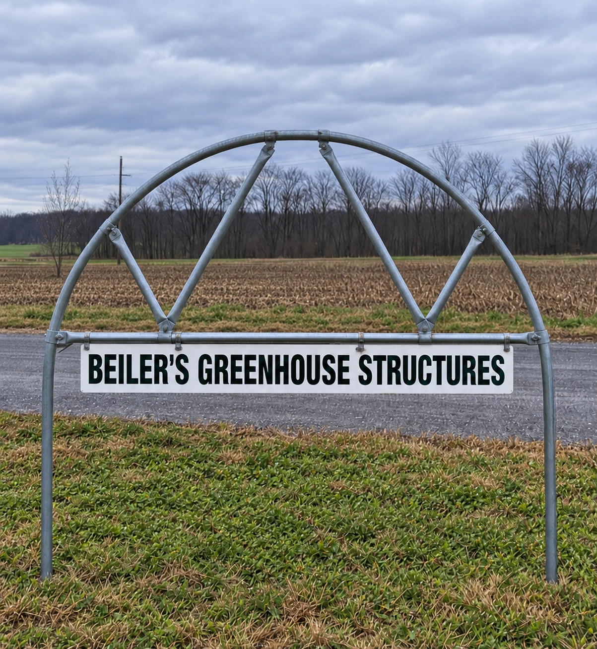 Beiler's Greenhouse Structures sign mounted on a steel frame beside a farm field.