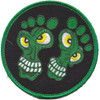 Circular embroidered patch with black field, green merrow border, and two cartoon green creature faces shaped like bare feet with yellow eyes and white teeth.