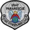 Badge-shaped embroidered patch with gray and silver field, black border, "USAF PARARESCUE" at top, angel with gold halo in red cloak kneeling over a blue and green globe, parachute canopy above, and scroll "That Others May Live" at bottom.