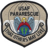 Badge-shaped embroidered patch in desert tan and khaki, black border, "USAF PARARESCUE" at top, angel with white wings in red cloak over globe, parachute canopy above, and "That Others May Live" scroll at bottom, all in subdued desert coloring.