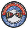 Round McMurdo Station Antarctica patch with white icicles over a red arc, station buildings and a snowy peak under falling snow, blue lower half and black merrowed border.