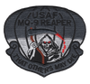 Badge-shaped subdued gray embroidered patch with "USAF MQ-9 REAPER" at top, a Grim Reaper with glowing red eyes holding a red scythe at center, parachute canopy behind, and "That Others May Die" scroll at bottom, in dark gray with red accents.