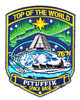 Shield-shaped embroidered patch for Pituffik Space Base showing the aurora borealis in green and cyan sweeping across a deep blue Arctic sky, the base pyramid radar dome in the center, a dogsled team on the ice below, a gold North Star at the top, and 76N in gold at lower right, with TOP OF THE WORLD arched above and PITUFFIK SPACE BASE on the lower banner.
