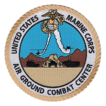 Round Marine Corps Air Ground Combat Center patch with an eagle, globe and anchor trailing smoke over two aircraft, black armored vehicles and brown desert mountains, inside a white lettered band and gold rope border.