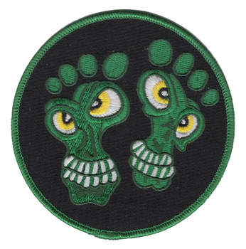 Circular embroidered patch with black field, green border, and two cartoon green creature faces shaped like bare feet with yellow eyes and white teeth.