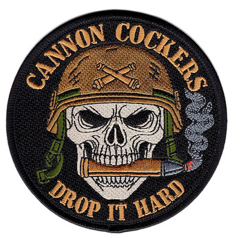 Round black embroidered patch with a skull in a steel pot helmet biting an artillery round, crossed shells on the helmet crown, gold text reading CANNON COCKERS across the top and DROP IT HARD across the bottom, merrowed border.