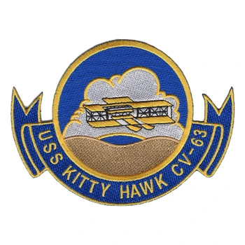 USS Kitty Hawk CV-63 patch with a gold and white Wright Flyer over silver clouds and a tan sand hill inside a blue circle, above a blue ribbon with gold lettering.