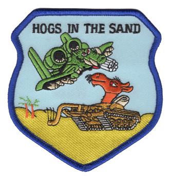 Shield-shaped embroidered patch with blue border, light blue sky background, and sandy desert floor. A cartoon green A-10 Warthog with oversized eyes dives from upper left while a red-orange camel emerges from a tan enemy tank below. "HOGS IN THE SAND" in black text across the top. Desert Storm A-10 Warthog morale patch.