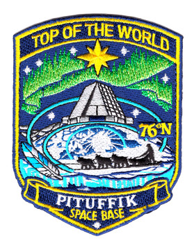 Shield-shaped embroidered patch for Pituffik Space Base showing the aurora borealis in green and cyan sweeping across a deep blue Arctic sky, the base pyramid radar dome in the center, a dogsled team on the ice below, a gold North Star at the top, and 76N in gold at lower right, with TOP OF THE WORLD arched above and PITUFFIK SPACE BASE on the lower banner.