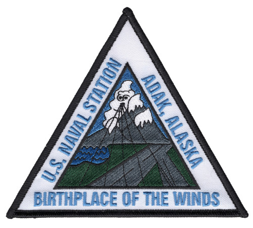 Triangular embroidered patch for U.S. Naval Station Adak Alaska featuring volcanic peaks, a white wind-spirit figure rising from the summit, and the motto Birthplace of the Winds along the base in blue lettering on a white field with black merrowed border.
