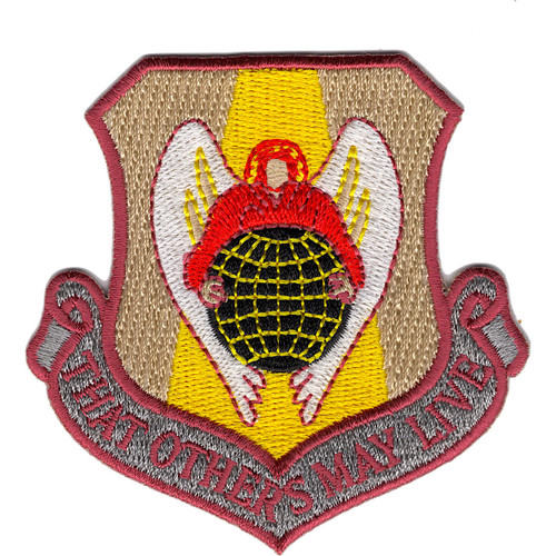 Shield-shaped embroidered patch in desert tan and khaki with maroon border, angel with white wings in red cloak over green globe on gold inner field, and "That Others May Live" scroll at bottom.