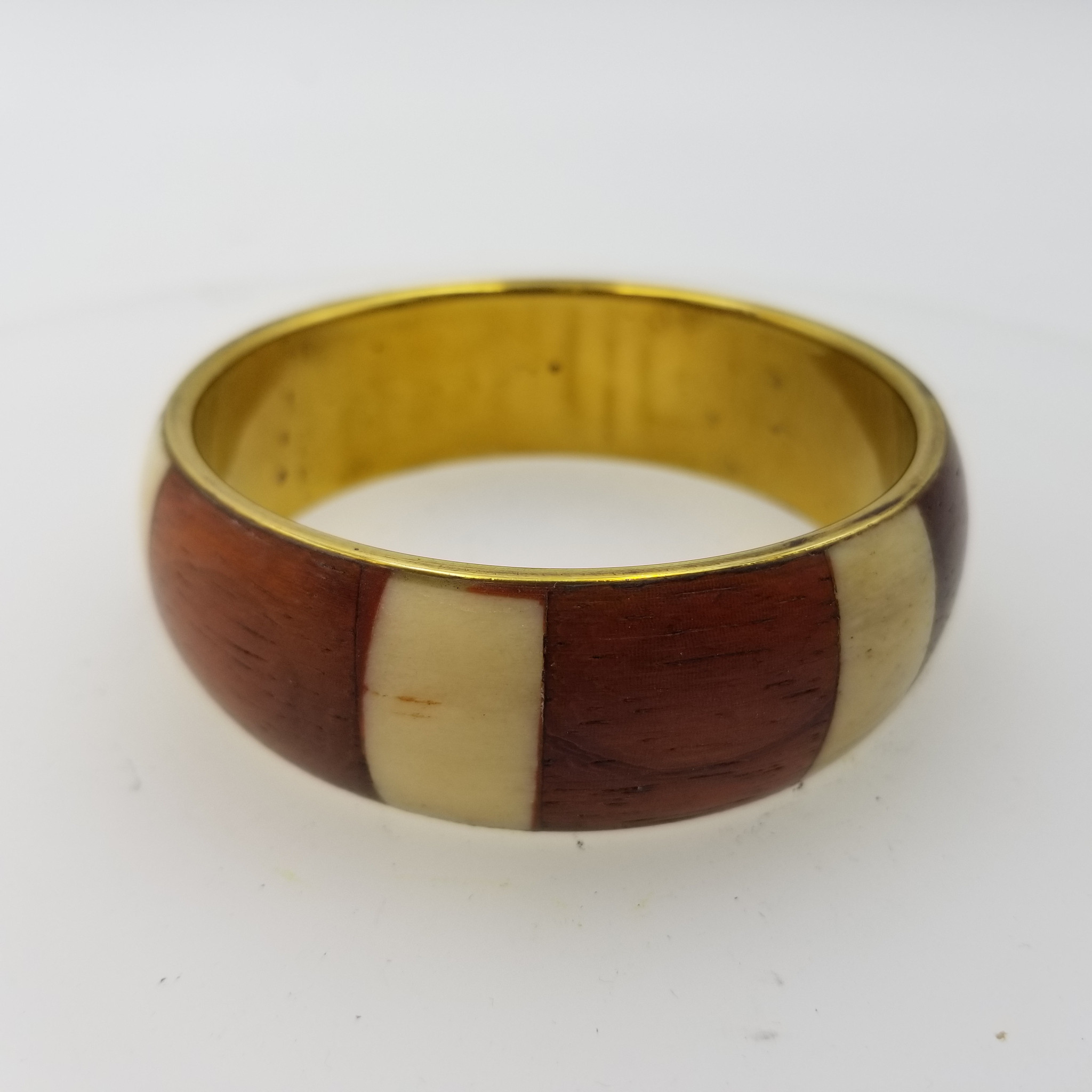 This is a vintage-style inlay bangle bracelet with a warm, classic look.

Description

Type: Rigid bangle bracelet with a smooth, rounded profile.

Materials/Look: Gold-tone metal interior with an outer band featuring alternating wood and ivory/cream inlay panels.

Design: The bracelet has a block/stripe pattern—rich reddish-brown woodgrain sections separated by lighter cream-colored panels for contrast.

Finish: Polished and glossy, with subtle natural grain visible in the darker sections.

Style/Vibe: Timeless and preppy-classic, with a slightly retro / vintage feel—easy to wear alone or stacked with other bangles.