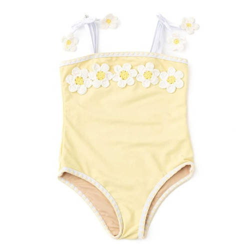 A butter-yellow Shade Critters Butter Daisy one-piece swimsuit is shown at an angle, featuring a row of white and yellow 3D crochet daisies across the chest and white straps with floral appliqués.